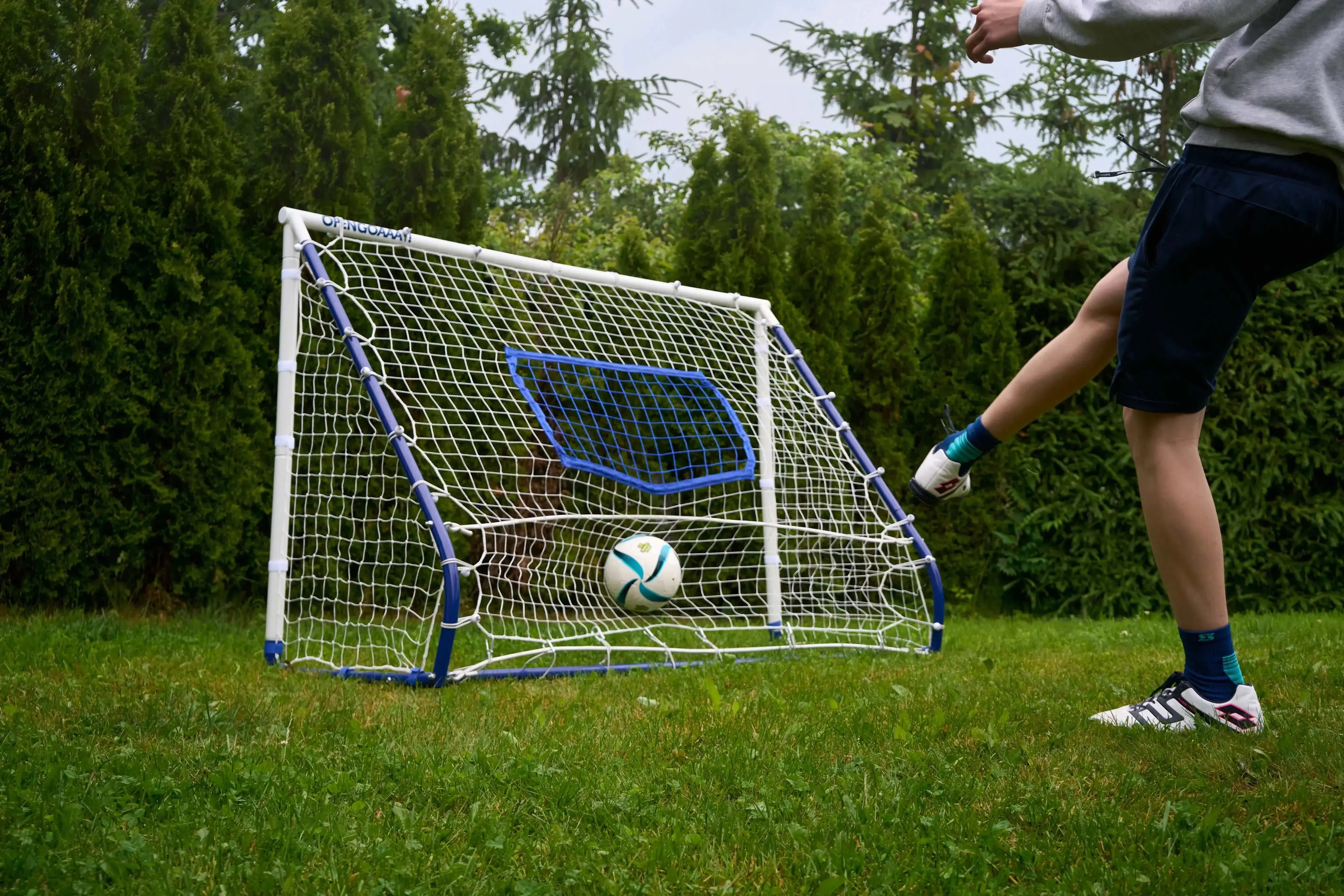 A person kicking a soccer ball toward a compact football rebounder goal trainer in a backyard setting.